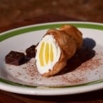 Dolce tipico siciliano in un piatto, con un paesaggio siciliano sullo sfondo.